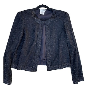 May include: A black, open-front jacket with a textured, beaded surface. The jacket has long sleeves and a rounded neckline. The interior lining is a darker shade of black. The jacket is hanging on a black hanger.