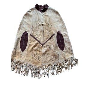 Puede incluir: Capa de ante beige con cuello y ribetes marrones. La capa presenta un diseño dibujado a mano de caballos y árboles, con flecos en el borde inferior. Dos bolsillos ovalados marrones están en la parte delantera. Prenda única de estilo bohemio.