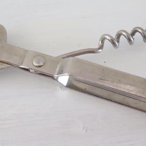 May include: A silver-colored metal multi-tool kitchen utensil featuring a corkscrew, a bottle opener, and a can opener blade. The tool has a hinged design with a long, flat handle and a sharp, curved metal point for piercing cans.