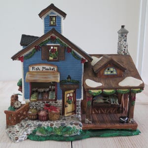 May include: A ceramic fish market building figurine featuring a blue exterior, a brown roof with snow accents, and a stone chimney. The front displays a sign reading Fish Market, baskets of produce, a yellow door, and a covered porch with fish.