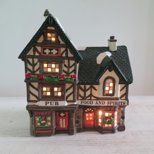 May include: A ceramic miniature building featuring a Tudor-style pub with timber framing, a dark shingled roof with snow accents, and illuminated windows. The facade displays the text PUB and FOOD AND SPIRITS, decorated with green wreaths and red bows.