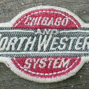 May include: An embroidered fabric patch featuring the text CHICAGO AND NORTH WESTERN SYSTEM. The design includes a central rectangular section with white lettering on a dark gray background, flanked by two semi-circular red sections with white borders.