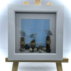 Puede incluir: Una obra de arte enmarcada con tres gaviotas azules posadas en postes de madera conectados por una cuerda. Las gaviotas están hechas de piedras pintadas y los postes de madera a la deriva. El fondo es un cielo azul claro. La obra de arte se exhibe en un pequeño caballete de madera.