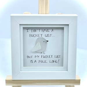 May include: A framed artwork with a white frame and a wooden easel. The artwork features a gray bird made of sea glass and the text "I DON'T HAVE A BUCKET LIST... BUT MY FUCKET LIST IS A MILE LONG!"
