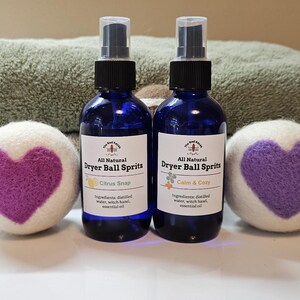 May include: Two blue glass spray bottles labeled "Dryer Ball Spritz" with "Citrus Snap" and "Calm & Cozy" scents. Two white dryer balls with purple heart designs are on either side. A green towel is in the background.