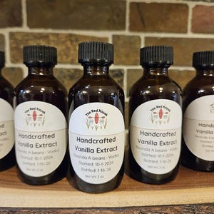 Op de afbeelding: Verschillende kleine, bruine glazen flessen met zwarte doppen. Elke fles heeft een wit etiket met de tekst "Handcrafted Vanilla Extract" en het logo "The Red Kayak". De etiketten bevatten ook details over de ingrediënten en de botteldatum. Elke fles bevat 59 ml.