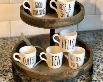 May include: A two-tiered wooden serving tray with a black metal stand. The tray is decorated with five white ceramic mugs with black lettering. The mugs read "Good Morning", "Rise & Shine", "Hello", "Tea", and "But First, Coffee".