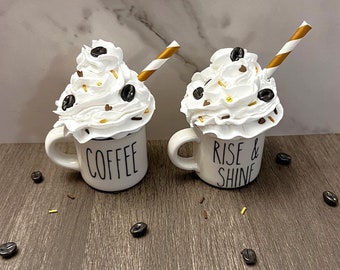 May include: Two white ceramic mugs with whipped cream, chocolate sprinkles, and coffee beans on top. One mug says "Coffee" and the other says "Rise & Shine".