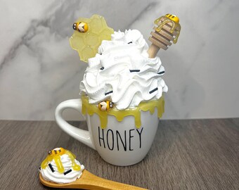 May include: A white mug with the word "HONEY" printed on it. The mug is filled with whipped cream, honey, and a small wooden honey dipper with a bee on top. There are also small bees on the side of the mug and on a small wooden spoon with whipped cream and honey.
