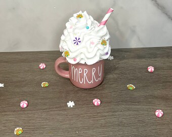 May include: A pink ceramic mug with the word "merry" on the side, filled with white whipped cream and topped with colorful sprinkles and a red and white striped straw.