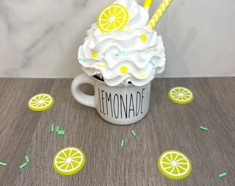 May include: White ceramic mug with the word "LEMONADE" printed on it. The mug is filled with whipped cream, yellow lemon slices, and yellow and blue sprinkles. Two yellow and white striped straws stick out of the whipped cream.