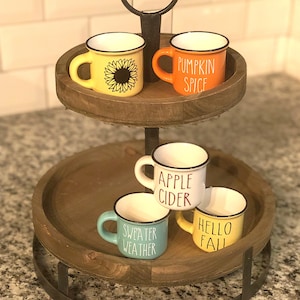 May include: A two-tiered wooden serving tray with a black metal stand. The top tier has two small mugs, one with a sunflower design and the other with the text "Pumpkin Spice". The bottom tier has four small mugs with the text "Apple Cider", "Sweater Weather", "Hello Fall", and a sunflower design.
