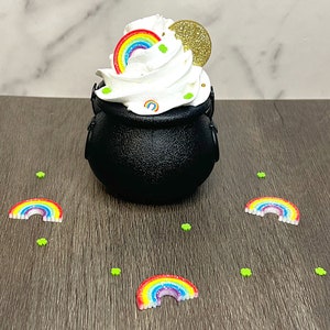 May include: A black cauldron with a white whipped cream topping decorated with a rainbow, a gold coin, and green shamrocks. The cauldron is surrounded by small rainbow and shamrock sprinkles on a wood surface.