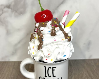 May include: White ceramic mug with a black rim and the words "ICE CREAM" printed on the front. The mug is filled with whipped cream, chocolate sauce, sprinkles, and a red cherry on top. Two striped straws stick out of the whipped cream.