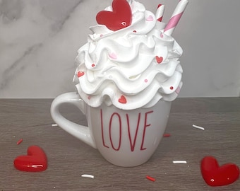 May include: White ceramic mug with the word "LOVE" printed on the front. The mug is filled with white whipped cream topped with red and pink heart sprinkles and a red heart candy. Two pink and white striped straws are sticking out of the whipped cream.