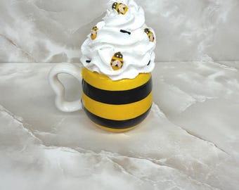 May include: A ceramic mug with a yellow and black striped design, topped with white whipped cream and bee-shaped decorations. The mug has a white handle and is set against a marble background.