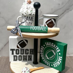 May include: A two-tiered decorative tray with football-themed items. Includes mugs, a rolling pin, and wooden blocks with football graphics and text. The tray is adorned with flags and a decorative spoon.