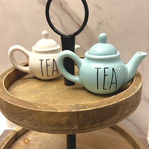 May include: Two miniature ceramic teapots, one white and one light blue, each with the word "TEA" in black lettering. They sit on a two-tiered wooden tray with a black metal handle. The teapots are decorative items.