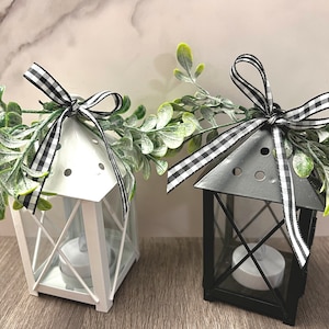 May include: Two small metal lanterns with black and white checkered ribbon bows and greenery. The lanterns are white and black and have a candle holder inside.