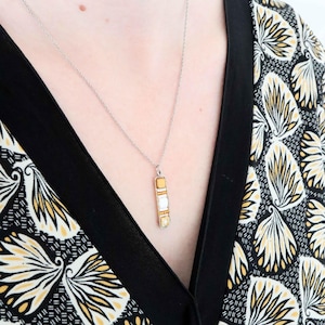 Könnte beinhalten: Silberkette mit rechteckigem Anhänger. Der Anhänger hat einen goldfarbenen Rahmen mit weißen und gelben Akzenten. Der Hintergrund zeigt ein schwarz-weiß gemustertes Oberteil mit gelben floralen Designs.