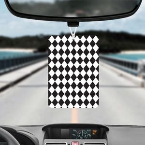 Puede incluir: Un ambientador rectangular para coche con un patrón de diamantes en blanco y negro, colgado de un espejo retrovisor. El fondo muestra una vista borrosa de una carretera, un puente y agua.