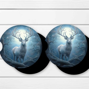 Puede incluir: Dos posavasos para coche con un majestuoso ciervo blanco de grandes cuernos, sobre un fondo de bosque místico. El diseño es predominantemente azul y blanco, con forma circular y base negra. Estos posavasos están diseñados para el portavasos del coche.