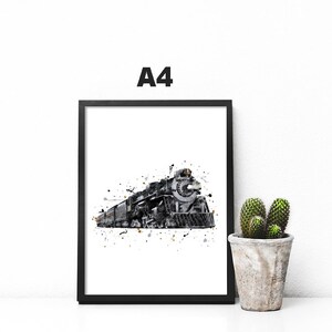 May include: A black and white watercolor print of a steam locomotive train in a black frame. The print is on a white background and has a splatter effect. The print is sitting on a white surface with a small cactus plant in a pot to the right.