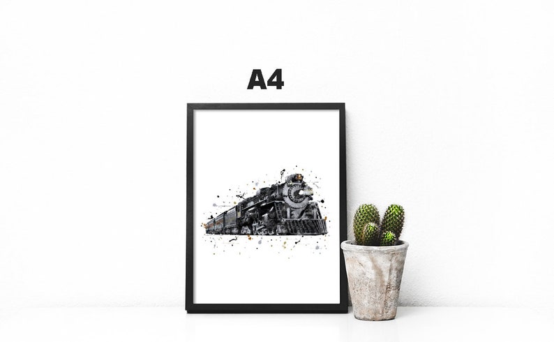 May include: A black and white watercolor print of a steam locomotive train in a black frame. The print is on a white background and has a splatter effect. The print is sitting on a white surface with a small cactus plant in a pot to the right.