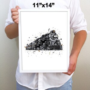 May include: A black and white watercolor print of a vintage steam locomotive train with gold and gray splatters on a white background. The print is framed in a white frame and measures 11 inches by 14 inches.