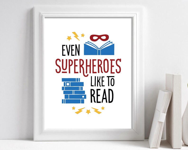 Puede incluir: Una impresi&oacute;n enmarcada en blanco con el texto "Incluso los superh&eacute;roes les gusta leer". La impresi&oacute;n presenta una pila de libros azules, una m&aacute;scara de superh&eacute;roe roja y rayos amarillos.