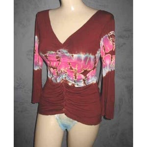 May include: A burgundy long-sleeve top with a V-neck and ruched detailing. The top features a tie-dye floral design in shades of pink, white, and blue across the chest and sleeves. The top is displayed on a mannequin.