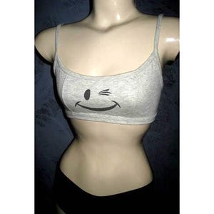 May include: A light gray crop top with thin straps and a winking smiley face graphic. The top is made of a soft material and is displayed on a mannequin. The bottom half of the mannequin is wearing black shorts.