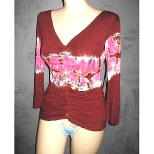 May include: A burgundy long-sleeve top with a V-neck and ruched detailing. The top features a floral pattern in shades of pink and white across the chest and upper sleeves. The sleeves are long and the fabric appears to be a soft, stretchy material.