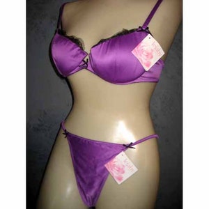 May include: A two-piece lingerie set in a vibrant purple hue. The bra features black lace trim and delicate straps, paired with a matching thong. Both pieces have small black bows and tags with a floral design.