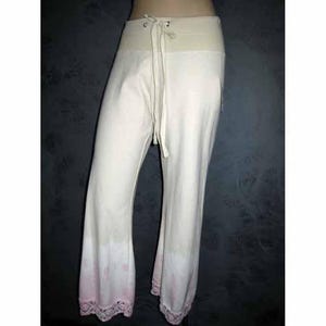 May include: Off-white lounge pants featuring a wide elastic waistband with a matching drawstring tie. The lower legs transition into a soft pink gradient pattern, finished with a delicate white lace trim along the bottom hem of each pant leg.