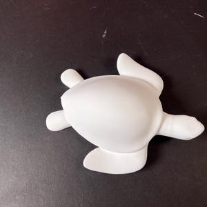 May include: A solid white sea turtle figurine with a smooth, matte finish. The turtle is depicted from a top-down perspective, showing its oval-shaped shell, four flippers extended outward, and a small head pointing towards the right side.