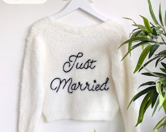 Könnte beinhalten: Ein weißer, flauschiger Langarmpullover mit dem in Schwarz gestickten Text Just Married auf der Vorderseite. Der Pullover hängt an einem weißen Kleiderbügel mit einer kleinen weißen Schleife vor einem schlichten weißen Hintergrund.
