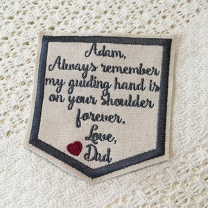May include: A gray and white embroidered patch with a heart and the text "Adam, Always remember my guiding hand is on your shoulder forever. Love, Dad".