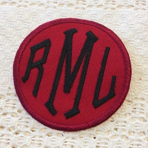 Peut inclure: Patch rond en tissu rouge avec un monogramme brodé noir "RML" en caractères gras.