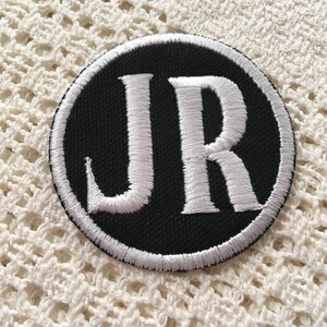 Pu&ograve; includere: Una toppa ricamata in bianco e nero con le lettere "JR" in un cerchio bianco su sfondo nero.