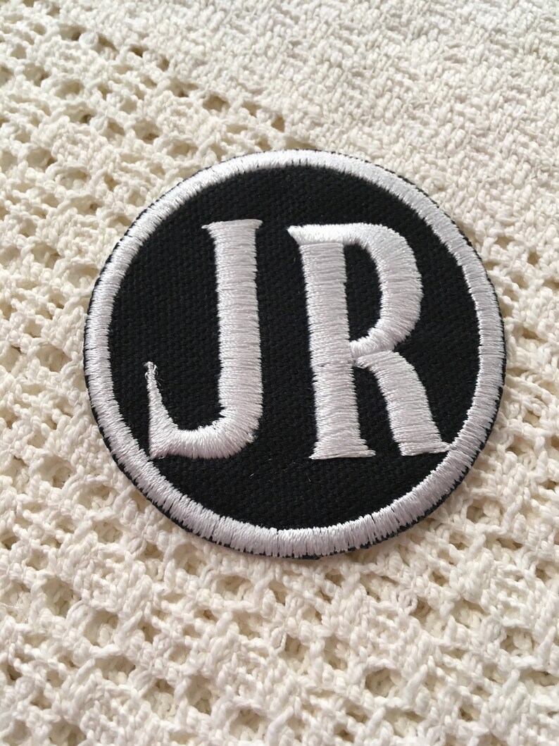 Pu&ograve; includere: Una toppa ricamata in bianco e nero con le lettere "JR" in un cerchio bianco su sfondo nero.