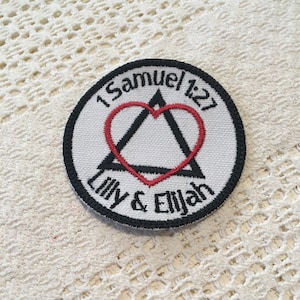 May include: A white round patch with black embroidery. The patch features a black triangle with a red heart inside. The text "1 Samuel 1:27" is above the triangle and the text "Lilly & Elijah" is below the triangle.