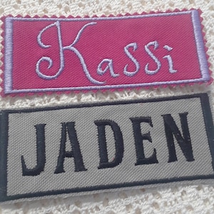 May include: Two fabric name tags. The top tag is pink with purple stitching and the name "Kassi" embroidered in purple thread. The bottom tag is gray with black stitching and the name "Jaden" embroidered in black thread.