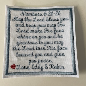 May include: White fabric square with blue embroidered border. The text reads "Numbers 6:24-26 May the Lord bless you and keep you may the Lord make His face shine on you and be gracious to you may the Lord turn His face toward you and give you peace, Love, Eddy & Robin".