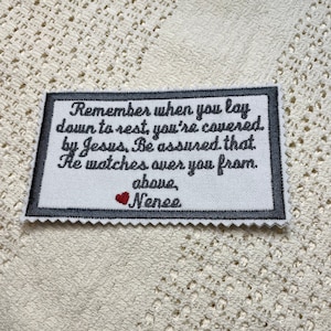 Może przedstawiać: Biały prostokąt z tkaniny z szarym obramowaniem i czarnym haftem z napisem "Remember when you lay down to rest, you're covered by Jesus. Be assured that He watches over you from above, Nenee". Małe czerwone serce jest wyszyte nad imieniem.