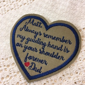 May include: A heart-shaped embroidered patch with a blue border. The patch is beige and features the text "Matt, Always remember my guiding hand is on your shoulder forever Dad" with a small red heart.
