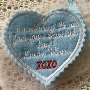 Puede incluir: Un corazón de fieltro azul claro con costuras blancas y el texto "Something Blue for your special day! Love, Mom XOXO" bordado en rojo.