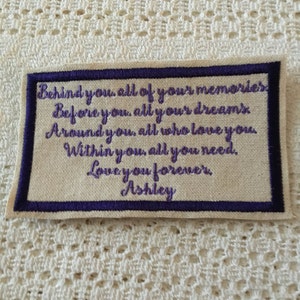 May include: A beige fabric patch with a purple embroidered message that reads "Behind you, all of your memories. Before you, all your dreams. Around you, all who love you. Within you, all you need. Love you forever. Ashley."