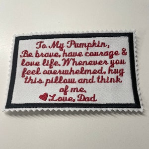 May include: A white fabric pillow with red embroidered text that reads "To My Pumpkin, Be brave, have courage & love life. Whenever you feel overwhelmed, hug this pillow and think of me. Love, Dad".