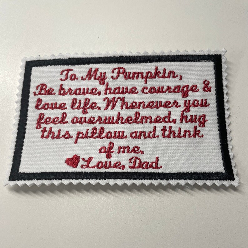 Op de afbeelding: Een wit stoffen kussen met rode geborduurde tekst die luidt "To My Pumpkin, Be brave, have courage & love life. Whenever you feel overwhelmed, hug this pillow and think of me. Love, Dad".